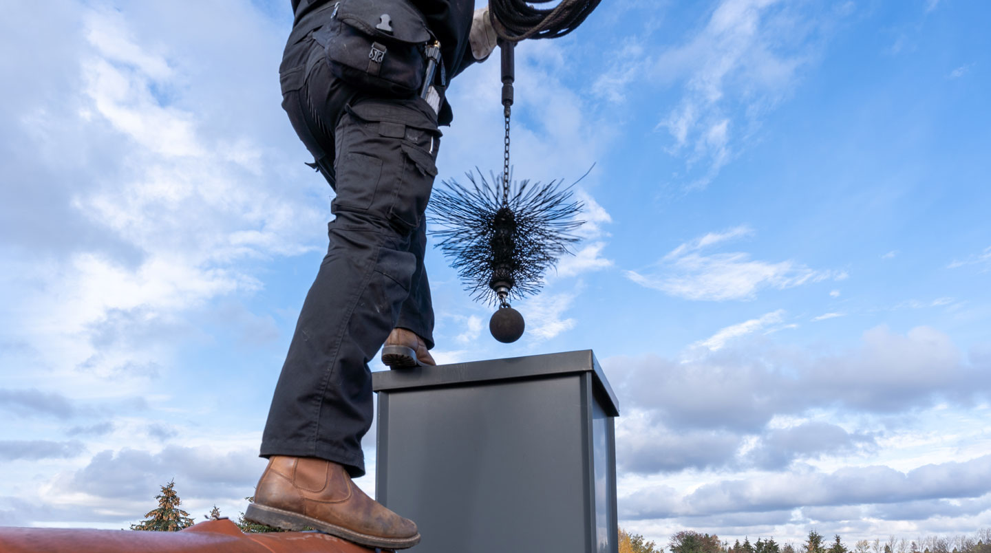 Ramoneur professionnel en vêtement de travail accroupi devant un poêle à bois, inspectant le conduit avec une lampe torche, outils visibles au sol